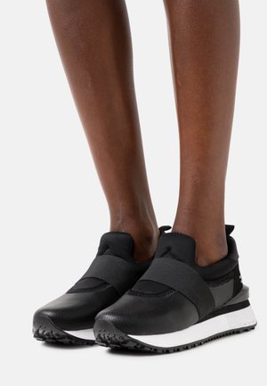 Paire de chaussures de sport noires à enfiler avec larges bandes élastiques portées sur des pieds à peau foncée, dotées de semelles texturées blanches et noires.