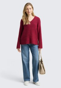 Bordowy sweter z dzianiny z dekoltem w serek, ściągaczem u dołu i długimi rękawami, w połączeniu z jasnoniebieskimi jeansami o prostych nogawkach oraz beżową torbą.