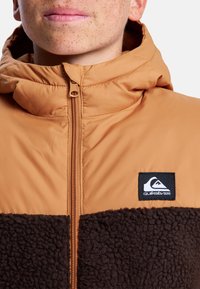 Brązowa i płowa kurtka na zamek z teksturą polarową. Posiada kaptur i naszywkę z logo na piersi. Detale obejmują przeszycia i design zamka.