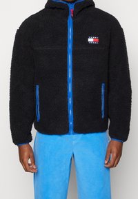 Svart fleecejacka med dragkedja fram, blå detaljer och sidofickor. Har en logomärken på bröstet och en huva.