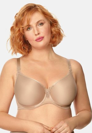 Kobieta o krótkich, kręconych rudych włosach, ubrana w beżowy, usztywniany biustonosz z koronkowymi detalami, pozująca na jednolitym jasnym tle.