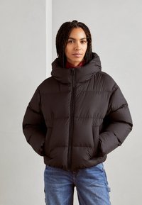 Schwarze Daunenjacke mit hohem Kragen, Reißverschluss vorne und gestepptem Muster. Weicher Stoff, lockere Passform, kombiniert mit blauen Jeans.