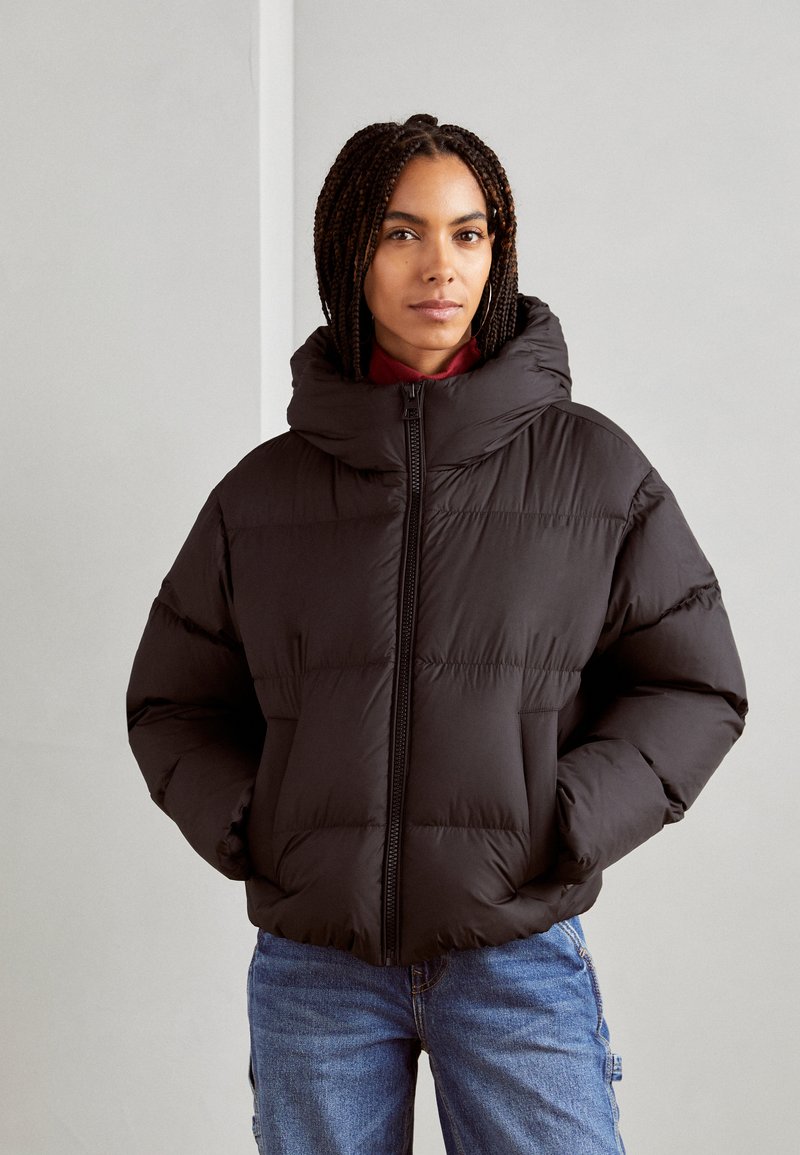 Schwarze Daunenjacke mit hohem Kragen, Reißverschluss vorne und gestepptem Muster. Weicher Stoff, lockere Passform, kombiniert mit blauen Jeans.