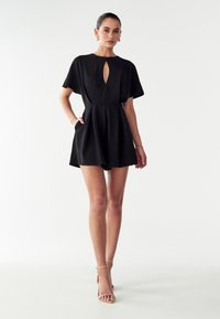 Črn kombinezon s kratkimi rokavi, izrezom v obliki ključavnice in dvema stranskima žepoma. Sametasta tekstura, oprijet pas in razširjeni kratki hlače.