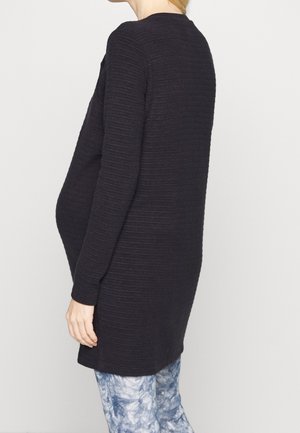 Frau, die ein schwarzes, strukturiertes Umstandsoberteil mit langen Ärmeln trägt, kombiniert mit blauen und weißen gemusterten Hosen, von der Seite gezeigt.