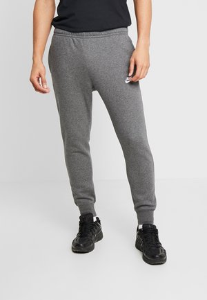 Mann trägt graue Nike-Sweatpants, schwarzes Shirt und schwarze Sportschuhe und steht vor einem einfarbigen Hintergrund.