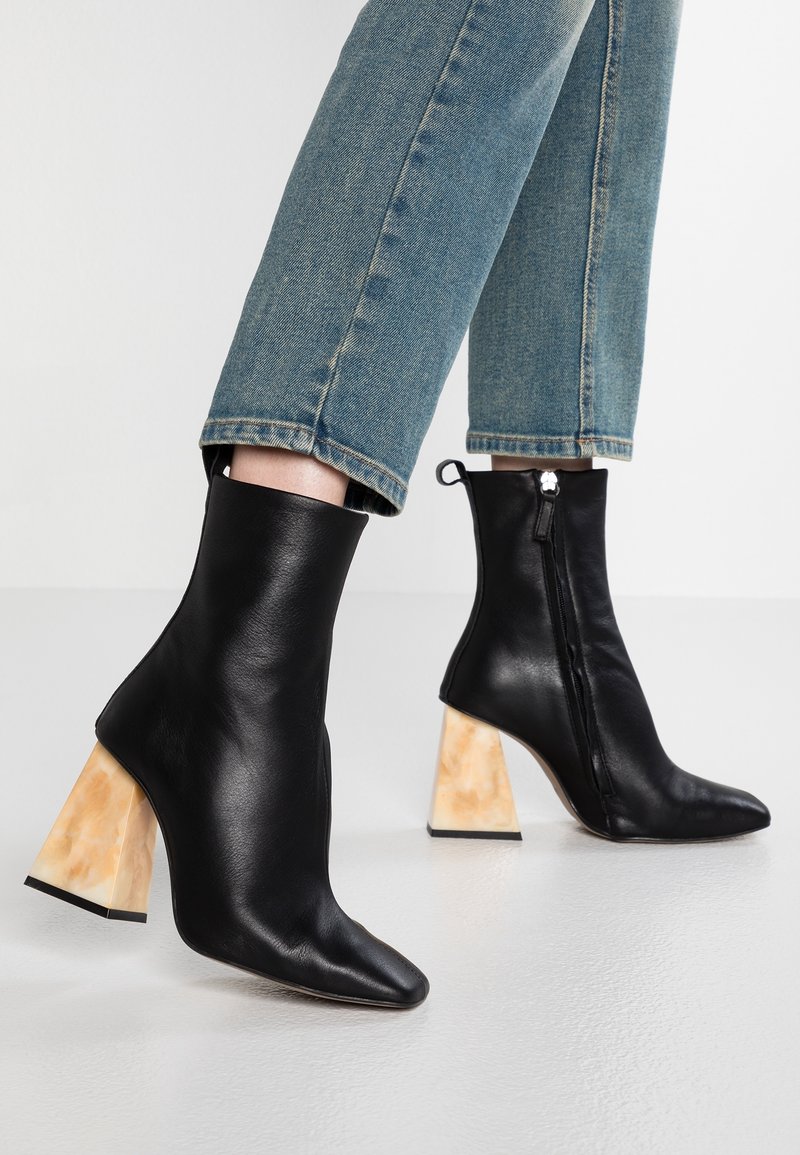 Bottines en cuir noir avec talons blocs beiges portées avec un jean en denim bleu, présentées sur un sol et un fond blancs.