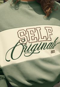 Groene en crèmekleurige sweatshirt met blokletters "SELF" en cursieve "Originals", voorzien van een zachte, textuurstof en geribbelde halslijn.