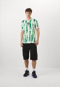 Chemise blanche à manches courtes avec des motifs de feuilles vertes, logo rond sur la poitrine. Associée à un short cargo noir et des sneakers noirs avec des accents bleus.