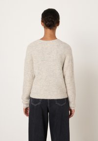 Sieviete ar tumšiem cirtainiem matiem, kas sakrauti zemu matu sasukā, nēsājot zelta lokveida auskarus, bēša krāsas ribotā džemperī un melnās platā gurnu biksēs, skatās prom no kameras.