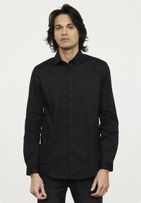 Chemise noire à manches longues avec un col structuré et une texture lisse. Équipée de boutons noirs et d'un design ajusté.