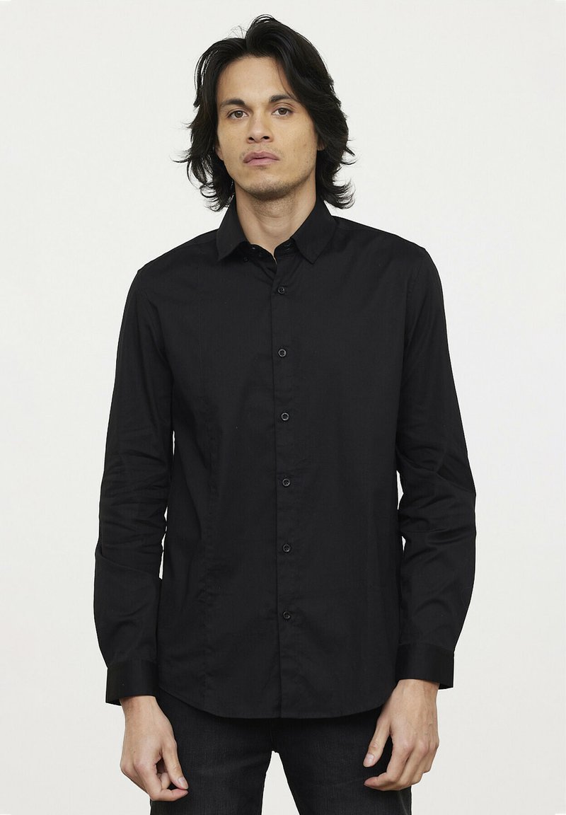 Chemise noire à manches longues avec un col structuré et une texture lisse. Équipée de boutons noirs et d'un design ajusté.