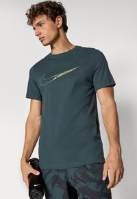 Temnozeleno bombažno majico s zlatim logotipom Nike swoosh, kratkimi rokavi, okroglim izrezom in sproščenim krojem, ki jo držite z črno plastenko za vodo.