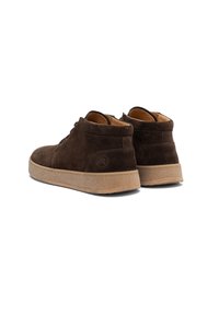 Une paire de bottines chelsea en suède marron foncé avec des semelles en caoutchouc beige, présentées de l'arrière sur fond blanc.