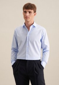Camicetta azzurra chiara con bottoni, realizzata in tessuto liscio, maniche lunghe e colletto a punta, abbinata a pantaloni scuri. Design semplice, senza fantasie.