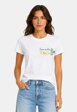 Donna con lunghi capelli castani che indossa una t-shirt bianca con testo in italiano e una piccola grafica di una bottiglia, abbinata a jeans azzurri chiari.