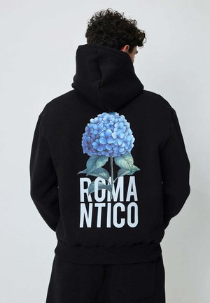 Homme portant un sweat à capuche noir avec une grande fleur d'hortensia bleue et le texte « ROMANTICO » au dos, tourné de dos sur un fond uni.