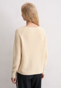 Gebreide trui in zacht beige, met een ribbeltextuur, ronde halslijn en lange mouwen. De zoom heeft zijsplitten voor meer bewegingsvrijheid.