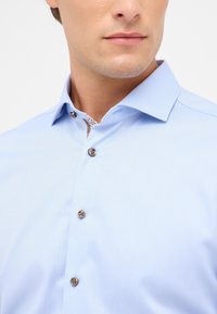 Camicia button-up azzurra in tessuto testurizzato, con bottoni marroni e un colletto interno a motivo contrastante.