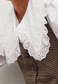 Col en dentelle blanche avec broderie florale et bords festonnés sur un gilet à motif pied-de-poule marron, doté de boutons foncés et d'une coupe ajustée.