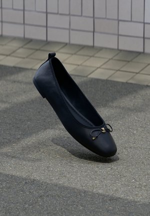 Sort balletflat med en lille sløjfe på toppen, svævende over en tekstureret grå fortov med en baggrund af flisevæg.