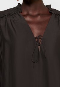 Blouse noire légère avec un col en V profond, épaules froncées et un cordon de serrage au niveau de l'encolure pour un ajustement personnalisable. Texture lisse.