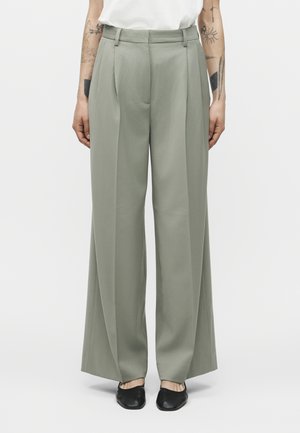 Άτομο που φοράει φαρδιά παντελόνια σε πράσινο χρώμα φασκόμηλου και μαύρα πλατφόρμες, με τατουάζ στους πήχεις, που στέκεται μπροστά σε απλό ανοιχτό φόντο.