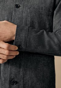 Tamno siva košulja s dugmićima sprijeda, džepom na prsima i manžetama s dugmićem. Materijal ima suptilnu teksturu i tkanu šaru.