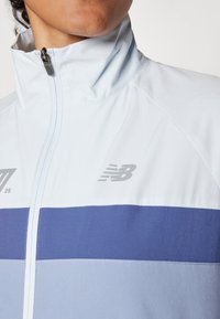 Giacca zip-up azzurro chiaro con colletto alto, caratterizzata da una striscia orizzontale blu scuro e un logo del marchio argentato sul petto. Texture liscia.