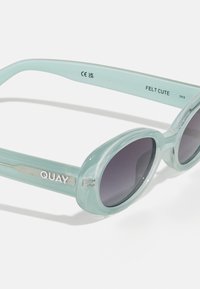Aqua atspalvių saulės akiniai su ovaliais tamsiai pilkais objektyvais, skaidriu rėmu ir metaliniu "QUAY" logotipu ant šonelio, pasižymintys išmaniu dizainu.