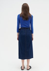 Mujer de pie de espaldas, vistiendo una blusa de manga larga azul, una falda midi de mezclilla oscura con bolsillos y zapatos negros tipo slip-on, contra un fondo liso.