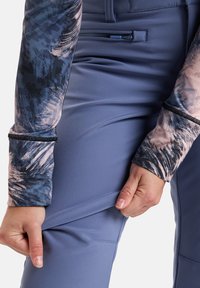 Langärmliges Oberteil mit mehrfarbigem abstraktem Muster, kombiniert mit schmalen blauen Hosen, die über eine Reißverschlusstasche und flache Nähte verfügen.