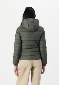 Dunjakke i mørkegrønn med horisontalt quiltet tekstur, hette, elastiske mansjetter og tettsittende silhuett, kombinert med beige bukser.