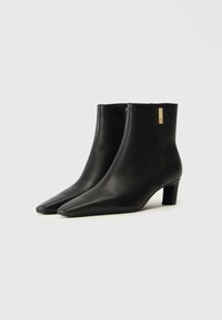 Sorte skinnankleboots med spiss tå og 3 cm blokkhel. Har et gullakcent på siden og en glatt tekstur.