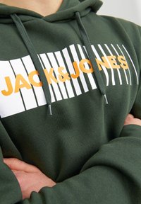 Felpa con cappuccio verde scuro con un design grafico bianco e arancione che include del testo. Presenta un cordino e polsini a coste. Materiale morbido e da indossare sopra.
