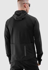 Zwarte sportjack met een capuchon, gestructureerde stof, een gecontroleerde achterzak met rits en reflecterende accenten. Heeft een getailleerd ontwerp en lange mouwen.