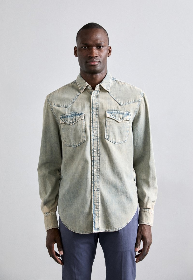 Camisa de mezclilla azul claro con dos bolsillos en el pecho, diseño con yugo puntiagudo, cierre de botones y mangas largas con puños abotonados.