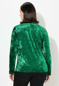 Top lungo a maniche verdi in velluto con una texture morbida, caratterizzato da un design aderente e un collo rotondo. La lucentezza uniforme esalta la ricchezza del tessuto.