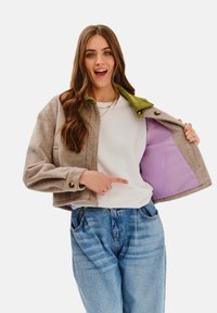 Skrócona beżowa kurtka z zielonym kołnierzem i fioletowym wnętrzem, noszona na białej koszuli, w zestawieniu z jasnoniebieskimi dżinsami.