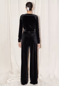 Jumpsuit nero in velluto con maniche lunghe, vita aderente e pantaloni a gamba larga. Texture liscia e design elegante creati per una vestibilità su misura.