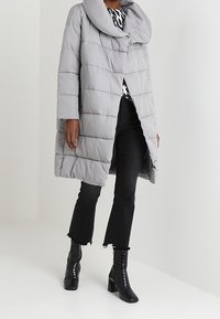 Světle šedá péřová bunda s velkým límcem, prošívaným designem, předními knoflíky a dlouhými rukávy, kombinovaná s černými rozšířenými džínami a botami na podpatku.