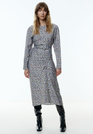 Langärmliges Blumenkleid aus leichtem Stoff, mit einer taillierten Taille, knielangem Rock und mehrfarbigem Blumenmuster. Getragen mit schwarzen Stiefeln.