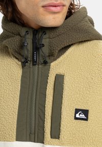 Fleece bunda v béžové a olivové zelené barvě se zipem vpředu. Má texturovanou tkaninu, nastavitelné šňůrky a vyšitý logotyp.