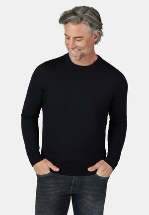 Uomo di mezza età con capelli grigi che indossa un maglione nero a maniche lunghe e jeans scuri, sorridente e con lo sguardo rivolto verso il basso, con le mani nelle tasche.