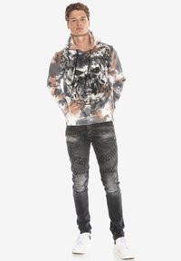 Kapuzensweatshirt mit einem Totenkopf-Graphic in einem grau, beige und weißen Batikmuster; kombiniert mit ausgewaschenen schwarzen Jeans mit strukturierten Akzenten.