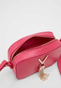 Pink textured leather crossbody bag with gold zipper and gold V-shaped tassel charm, partially open showing interior pocket.
