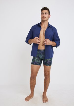 Marineblaues Hemd mit Knöpfen, untucked, kombiniert mit gemusterten Bade Shorts, die grüne und blaue Blätter auf einem dunklen Hintergrund zeigen. Barfuß.