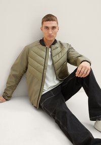 Ελαιόλαδο πράσινο μαξιλαρωτό μπουφάν με καπιτονέ σχέδιο ζιγκ-ζαγκ, γιακά που στέκεται, και μαύρο φινίρισμα. Φοριέται πάνω από ένα ανοιχτόχρωμο πουκάμισο και συνδυάζεται με σκούρα τζιν.