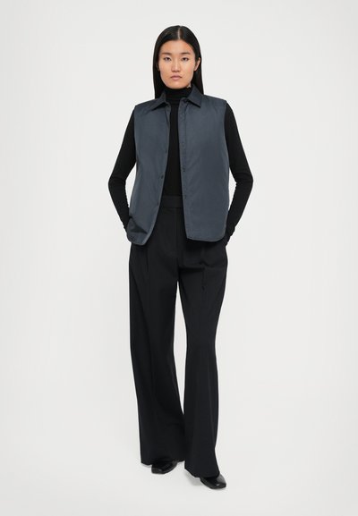 Mørk grå ermeløs vest med krage og knapper, båret over en svart turtleneck, kombinert med høy midje svarte ruter bukser.