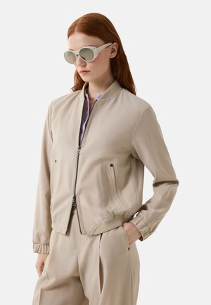 Beige Bomberjacke mit Reißverschluss, gerippten Bündchen und zwei Seitentaschen. Kombiniert mit hellgrauen, runden Sonnenbrillen. Glatte Textur.